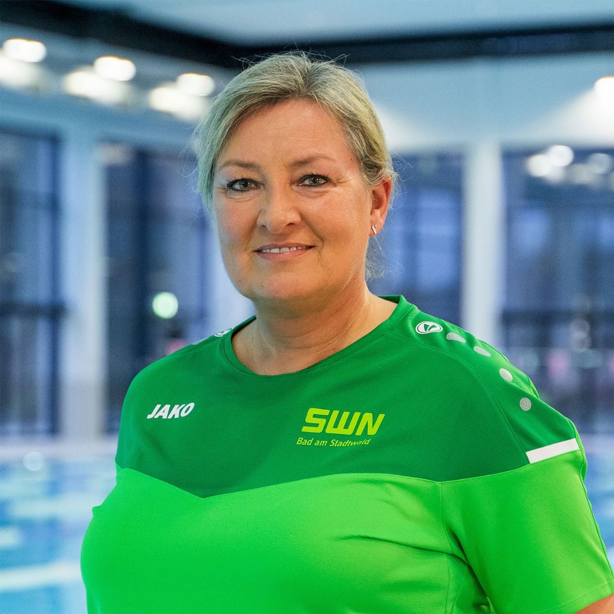 Eine Mitarbeiterin des Bad am Stadtwald (SWN) steht in grüner Dienstkleidung mit Logo vor dem Schwimmbecken. Sie blickt freundlich in die Kamera – der moderne Hallenbad-Bereich mit großen Fenstern ist im Hintergrund zu sehen.