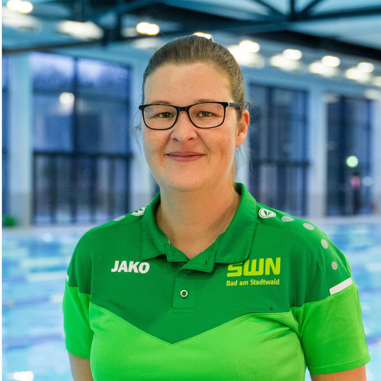 Eine Mitarbeiterin des Bad am Stadtwald (SWN) steht in grüner Dienstkleidung mit SWN-Logo vor dem Schwimmbecken. Der lichtdurchflutete Innenbereich des Hallenbads ist im Hintergrund zu sehen.