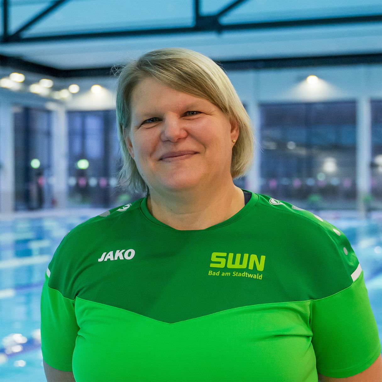 Eine Mitarbeiterin des Bad am Stadtwald (SWN) steht lächelnd in grüner Dienstkleidung mit Logo vor dem Schwimmbecken. Der moderne Hallenbad-Bereich mit großen Fenstern bildet den Hintergrund.
