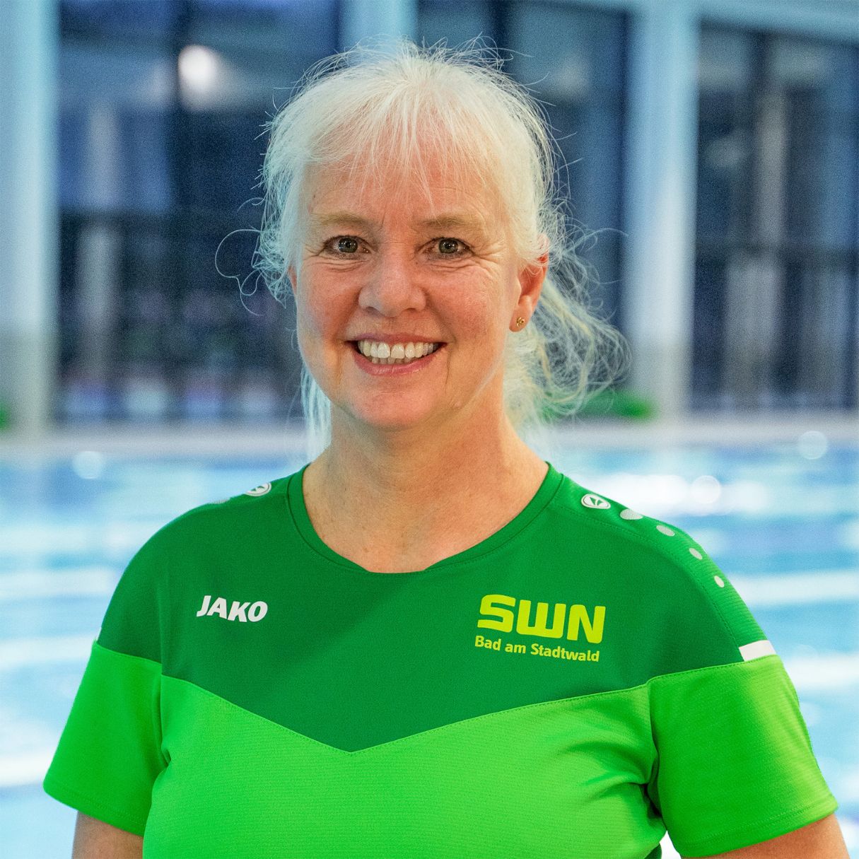 Eine Mitarbeiterin des Bad am Stadtwald (SWN) steht freundlich lächelnd in grüner SWN-Dienstkleidung vor dem Schwimmbecken. Im Hintergrund ist der helle Innenbereich des Hallenbads mit großen Fenstern und Wasserfläche zu sehen.