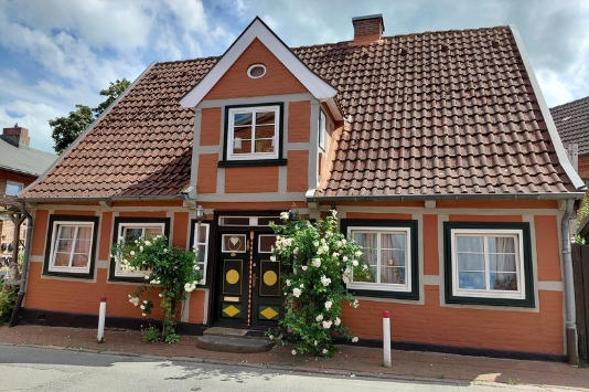 Historisches Fachwerkhaus mit roter Fassade, Satteldach, weißen Fensterrahmen und blühenden Rosensträuchern an der Straße
