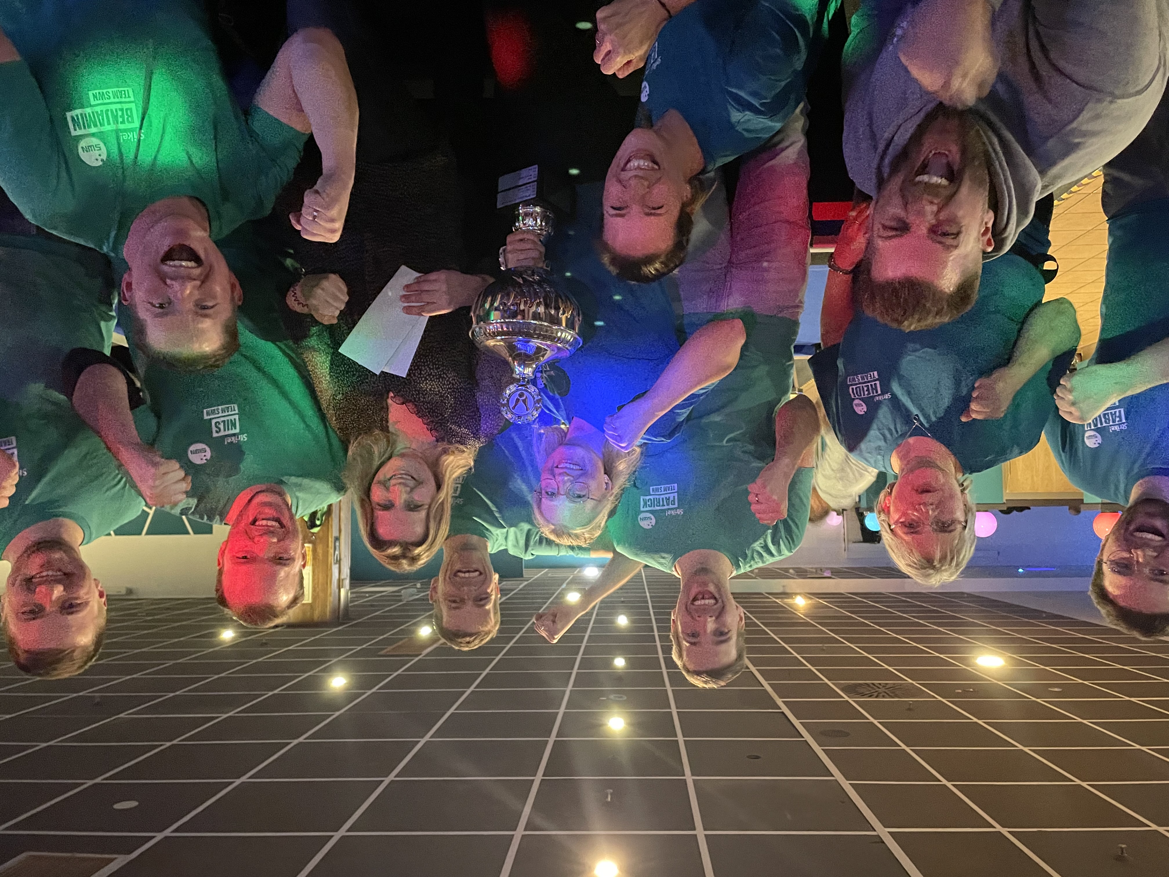 Ein Team jubelt gemeinsam bei einem Bowlingturnier, eine Frau in der Mitte hält einen großen Pokal in den Händen.