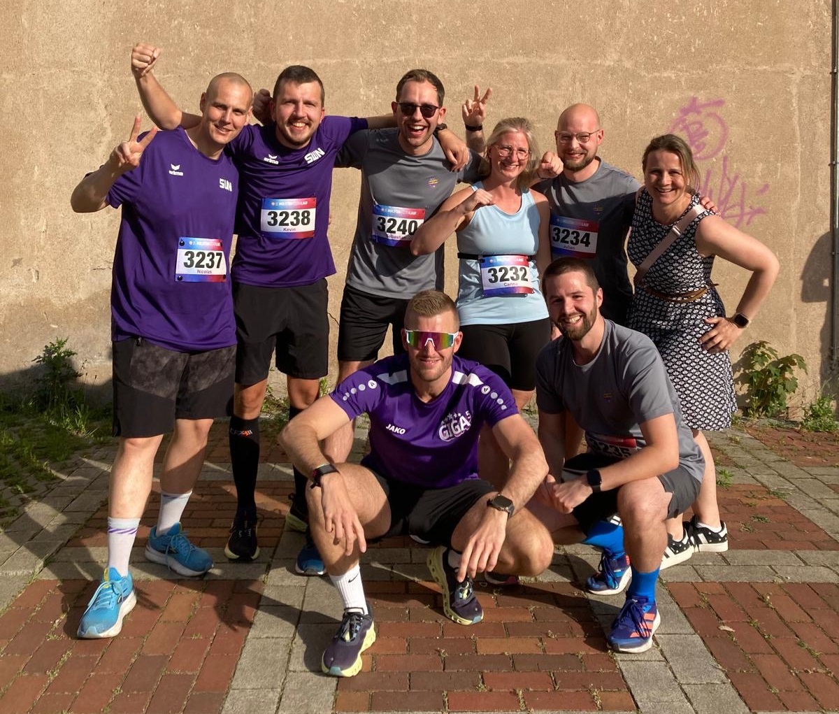 ine Gruppe von Läuferinnen und Läufern mit Startnummern posiert lachend nach einem Lauf vor einer Steinwand.