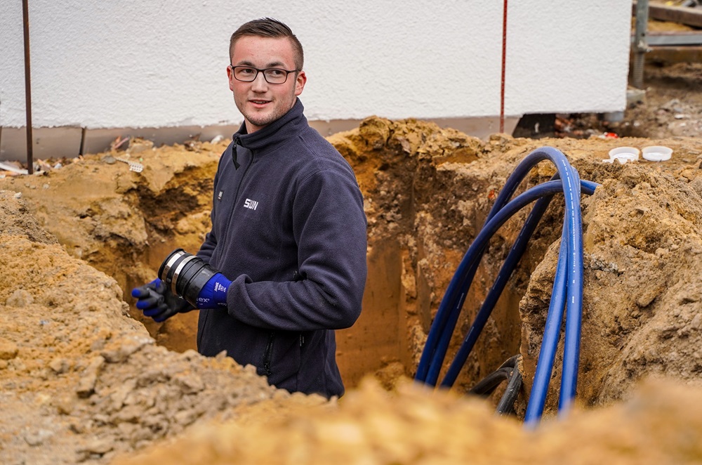 Mitarbeiter in SWN-Arbeitskleidung steht in einer Baugrube neben blauen Rohrleitungen und hält ein Verbindungsteil in der Hand.