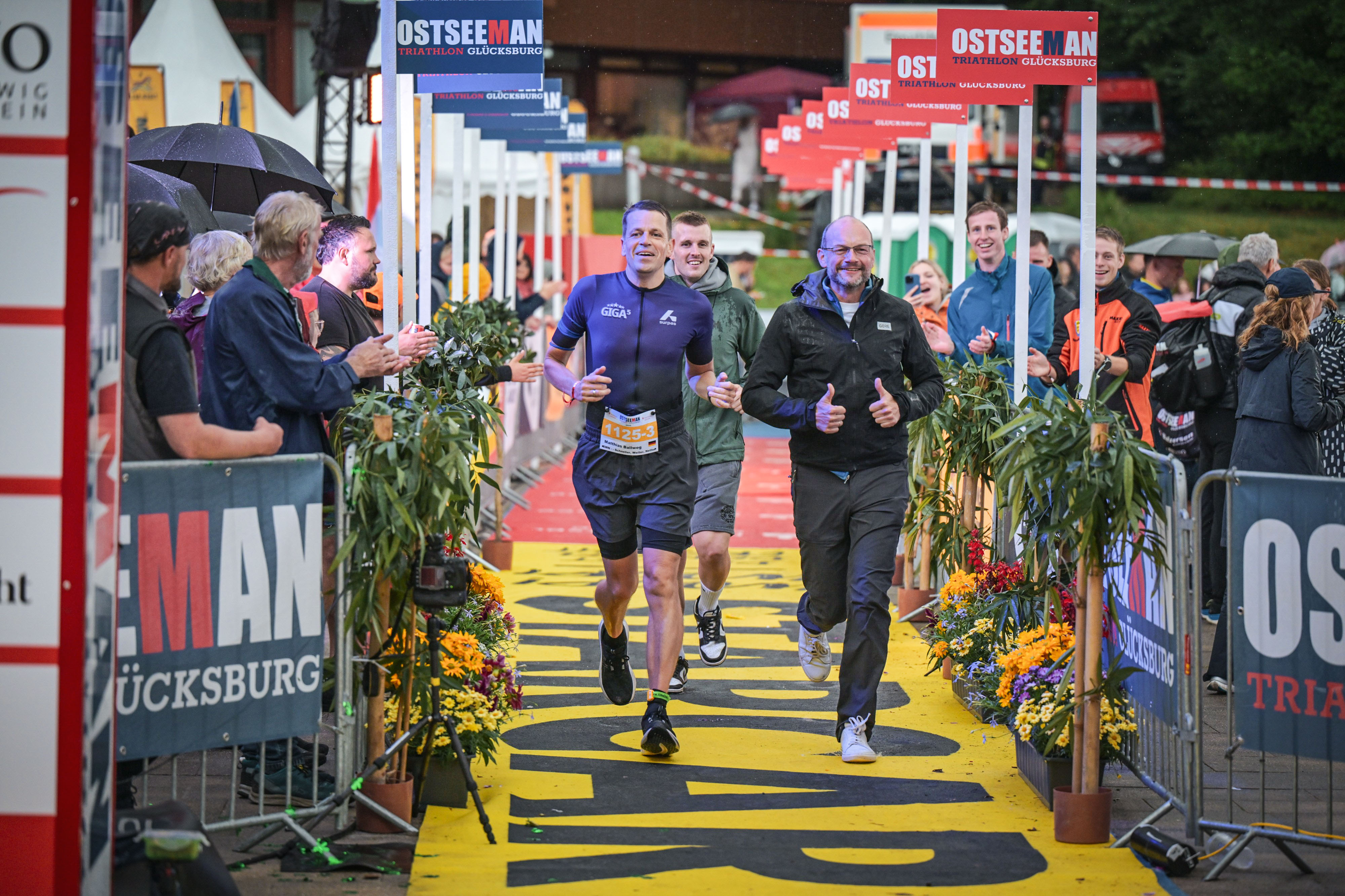 eilnehmende des OstseeMan Triathlons laufen gemeinsam durch den Zieleinlauf, während Zuschauer am Rand applaudieren.