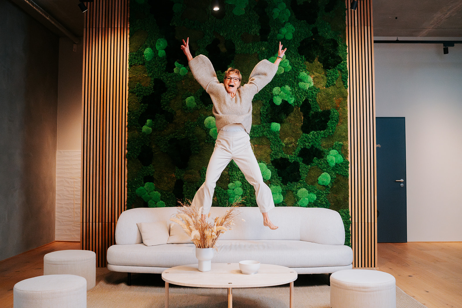 Eine lebendige Szene bei den Stadtwerken Neumünster (SWN): Eine Person springt voller Freude auf einem Sofa in einem modern gestalteten Raum mit einer grünen Mooswand im Hintergrund. Die energiegeladene Atmosphäre spiegelt Innovationsgeist und Dynamik wider.